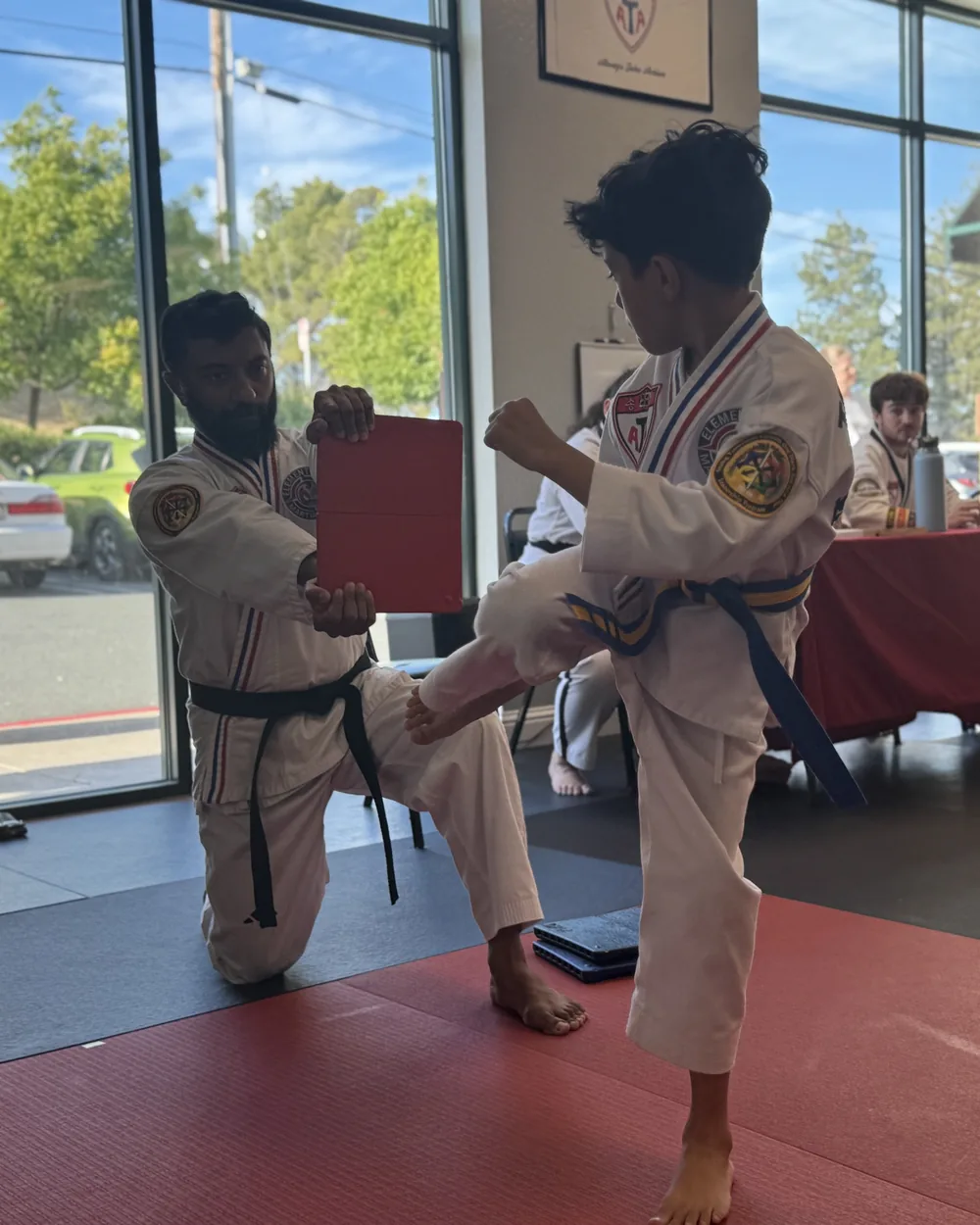 Black belt instructor kneeling to hold a breaking board for a young student mid-kick at Element ATA Martial Arts in Martinez CA